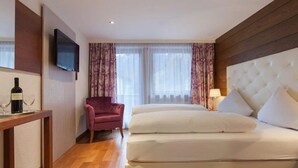 A hotel room with a large bed, a TV mounted on the wall, a chair, and a table with a bottle and glasses.