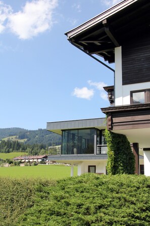 A modern house with a large glass window and a balcony, surrounded by greenery and a clear sky. - Pension Franglhof (Kirchberg in Tirol)