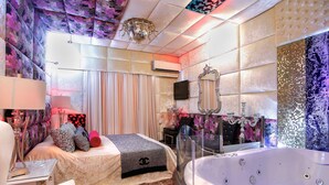 A luxurious bedroom with a large bed, a vanity with a mirror, a chandelier, and a bathtub. - Priamos - Adults Only (Athens)