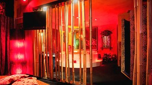 A room with a red-lit ambiance, featuring a bed with patterned bedding, a television mounted on the wall, and a decorative mirror. - Priamos - Adults Only (Athens)