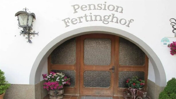 A building entrance with a sign reading "Pension Fertinghof," featuring a wooden door with glass panels, a wrought iron lamp, and potted plants. - Fertinghof (OBERAU)