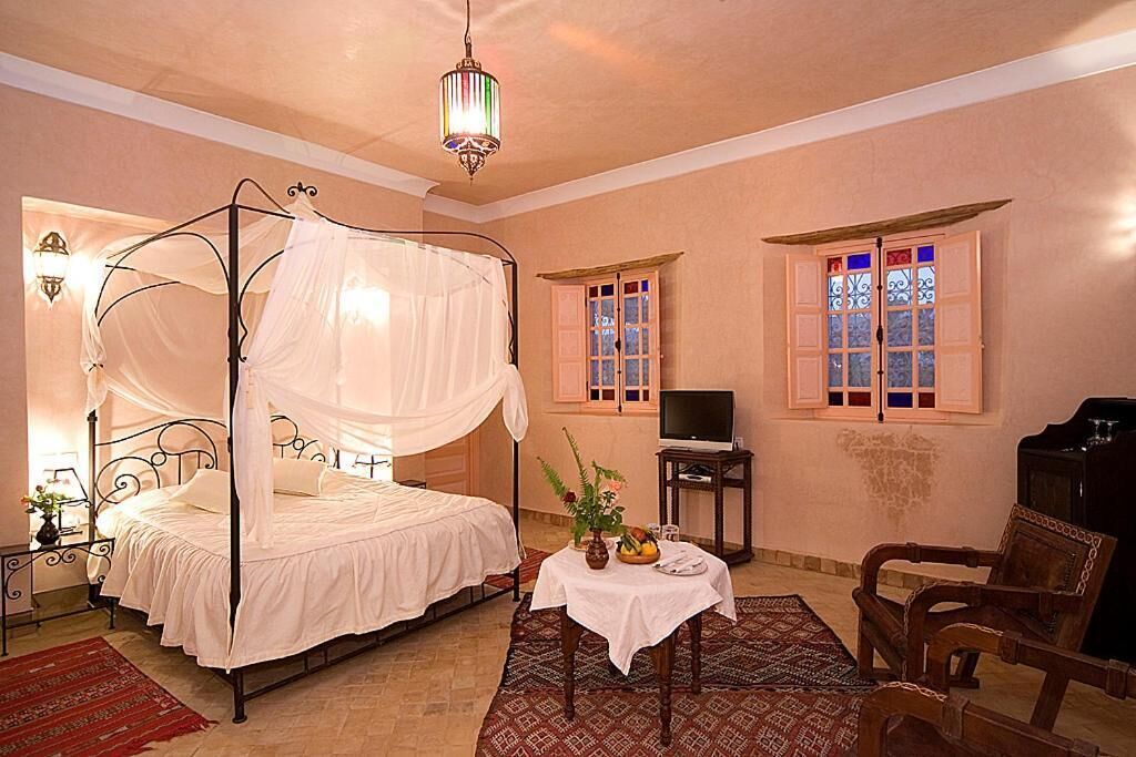 Una habitación con cama con dosel, un televisor sobre un mueble bajo, una mesita con un jarrón y fruta, y una silla.