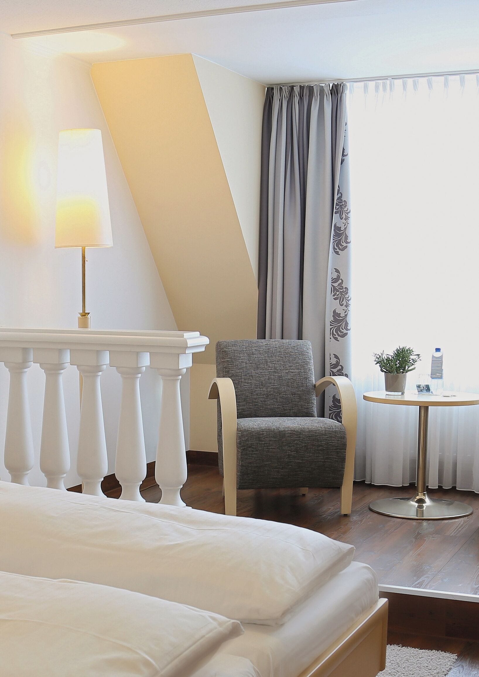 A hotel room with a bed, a chair, a small table with a plant and a bottle, and a window with curtains.