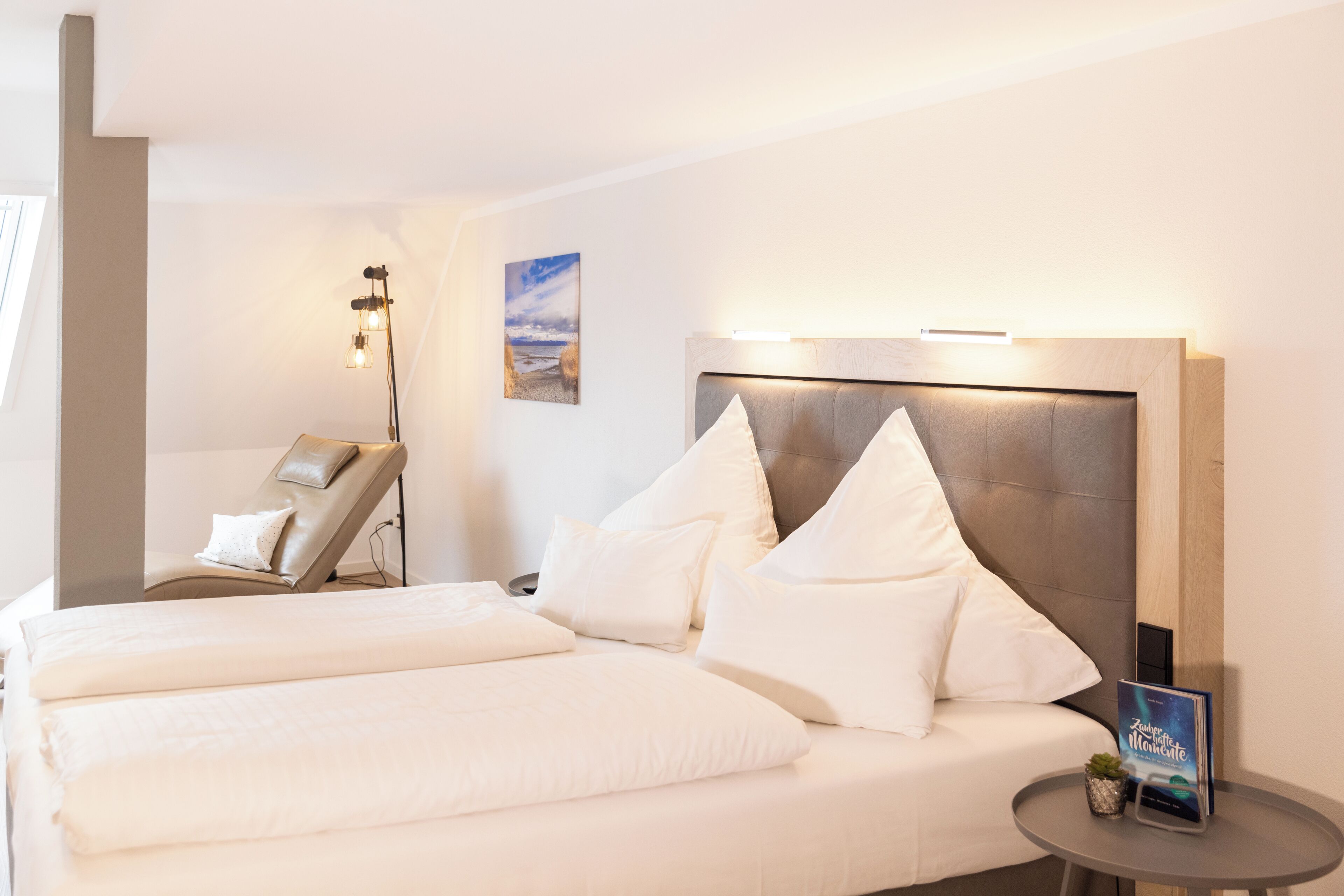 Une chambre d’hôtel avec un grand lit, un fauteuil massant, une petite table avec une plante et un luminaire fixé au mur.