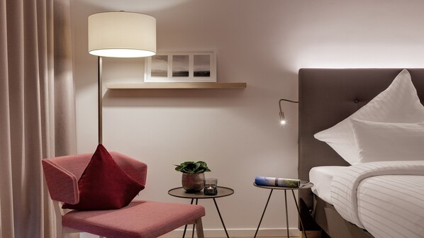 Habitación de hotel con cama, una silla roja, una lámpara de pie, una mesita con una planta y un vaso, y una estantería con marcos de fotos.
