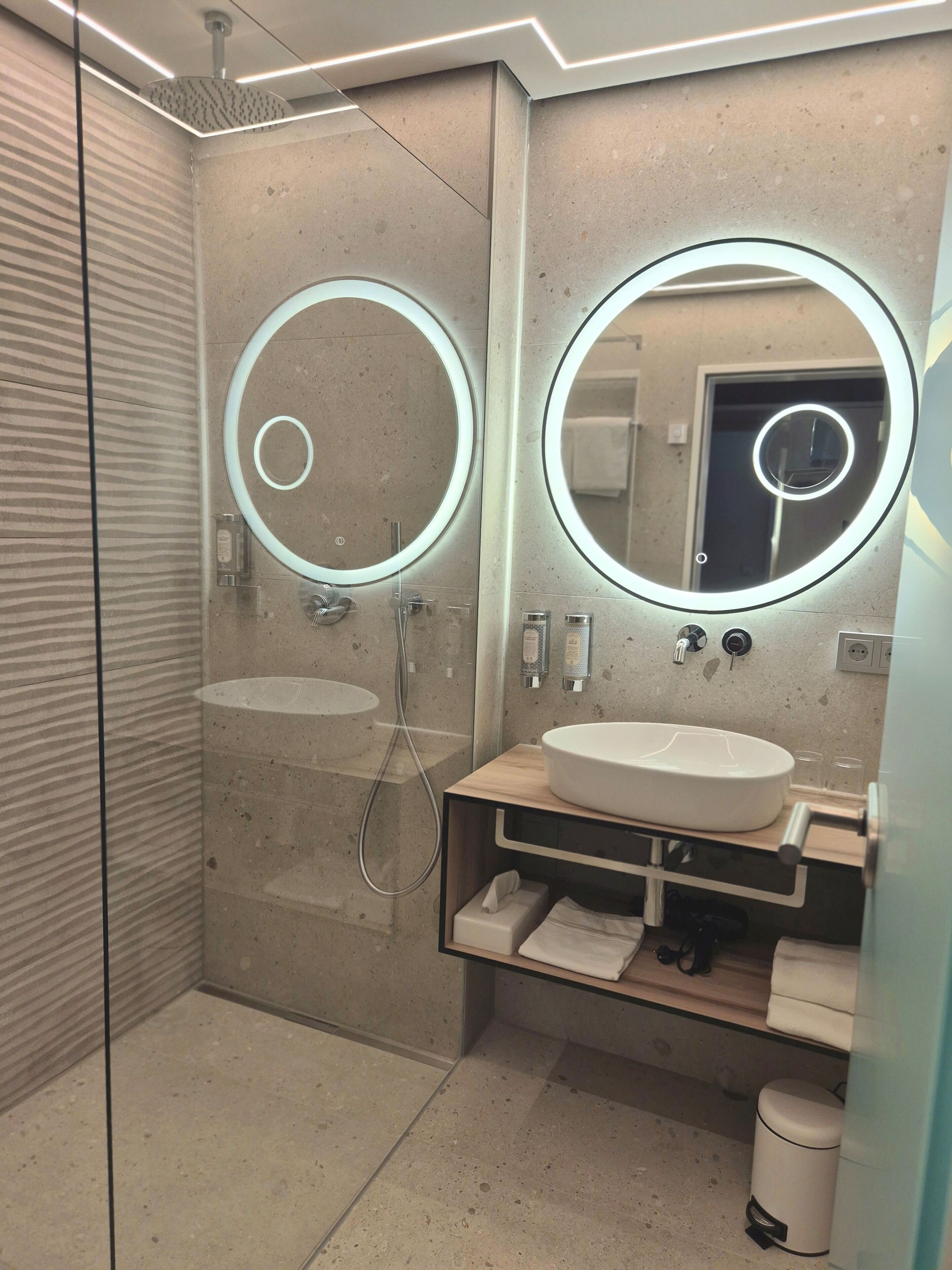 A modern bathroom with a glass-enclosed shower, a wooden vanity with a white sink, and two illuminated mirrors.