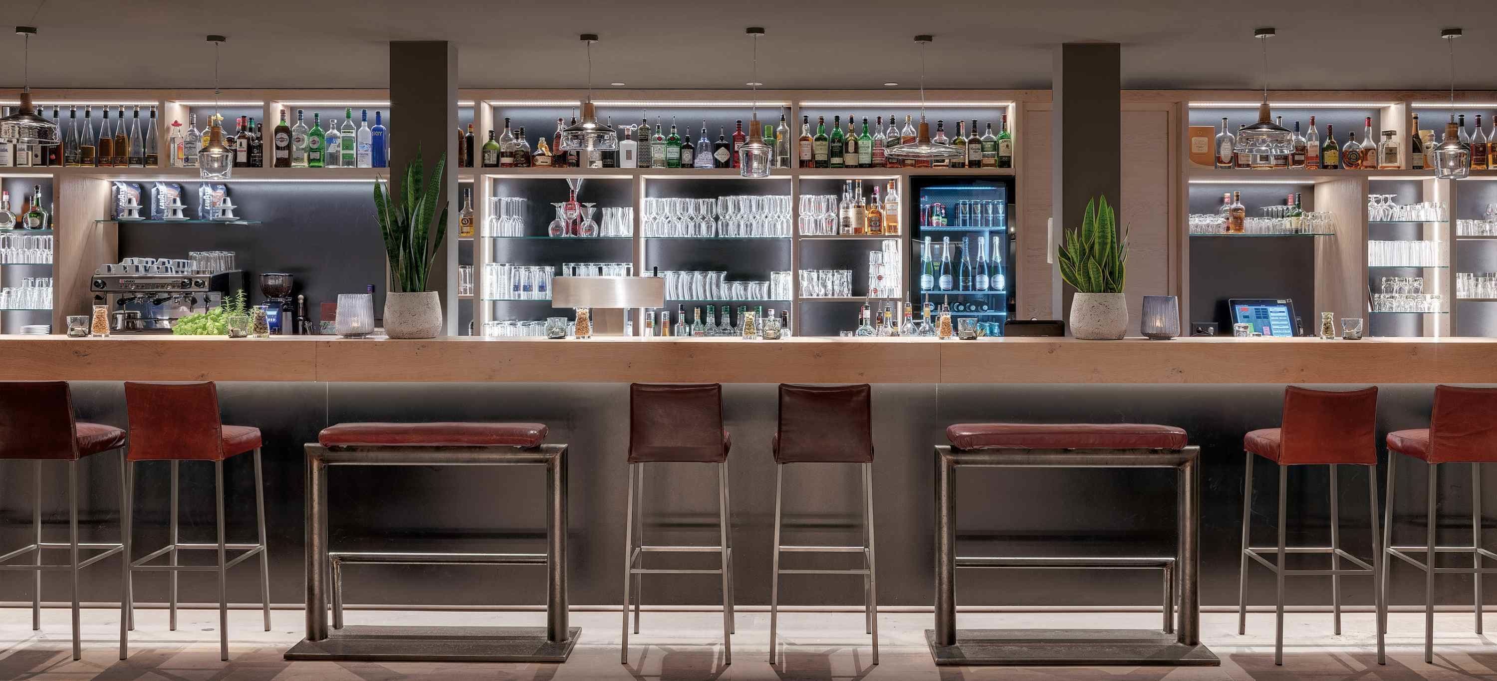 A bar area with multiple shelves of bottles and glasses, a bar counter, and four stools.
