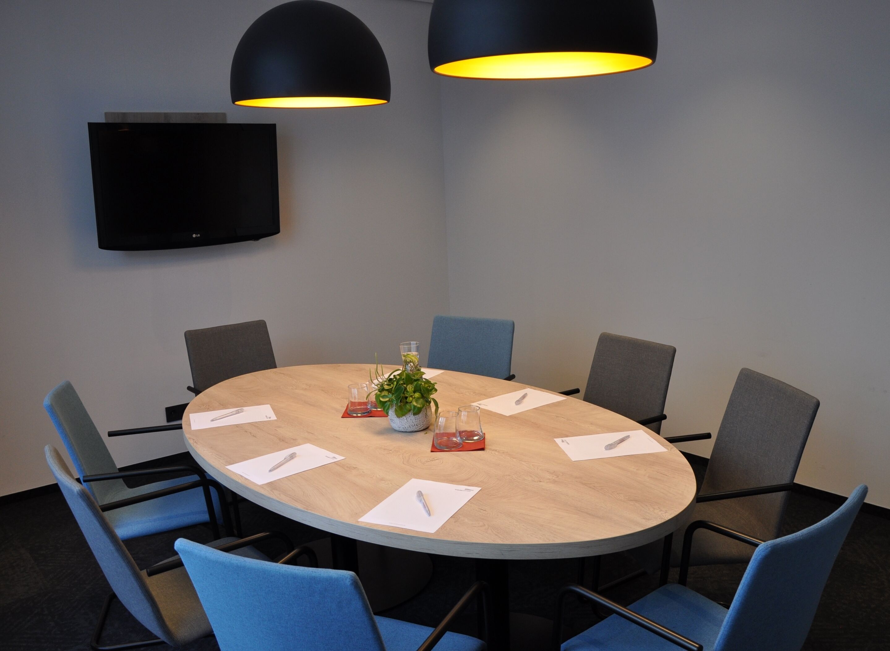 Een vergaderruimte met een ronde tafel, blauwe stoelen, een potplant en twee zwarte hanglampen.