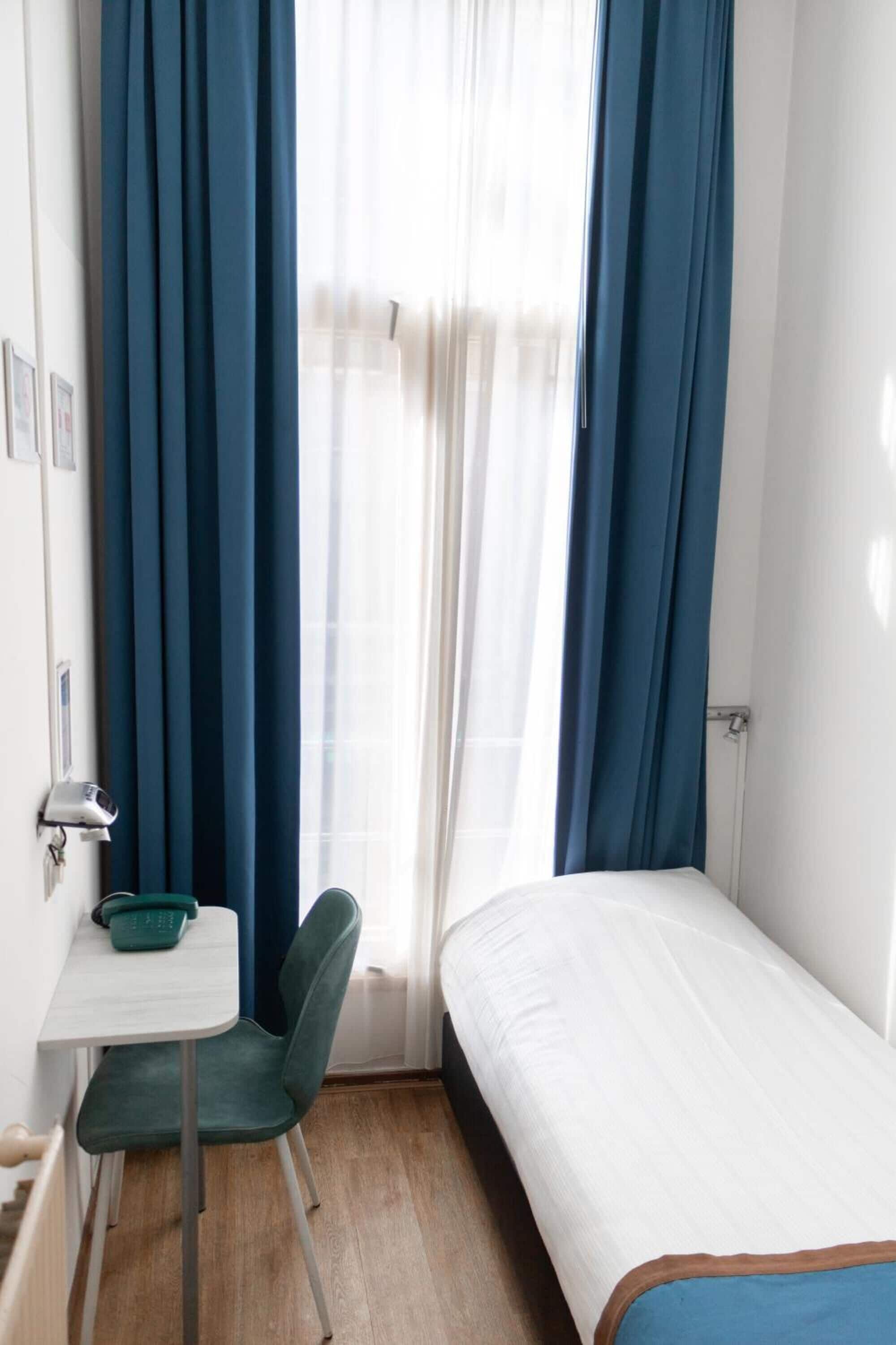 A small, single-bed room with a desk and chair, a window with blue curtains, and a white wall.
