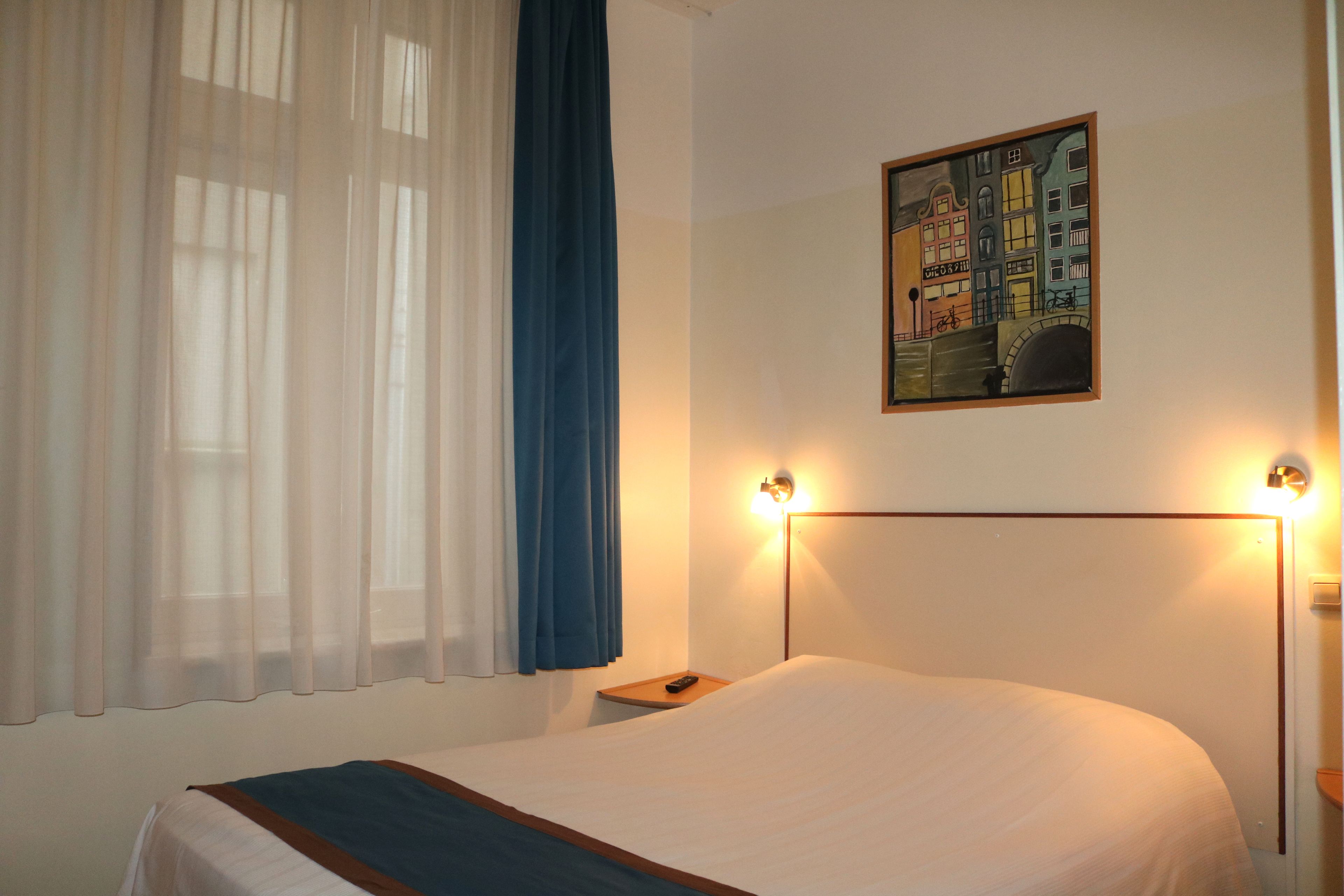A neatly made bed with a white and blue bedspread, a nightstand with a remote control, and a painting of a cityscape on the wall.
