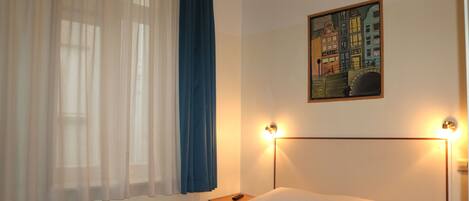 A neatly made bed with a white and blue bedspread, a nightstand with a remote control, and a painting of a cityscape on the wall.