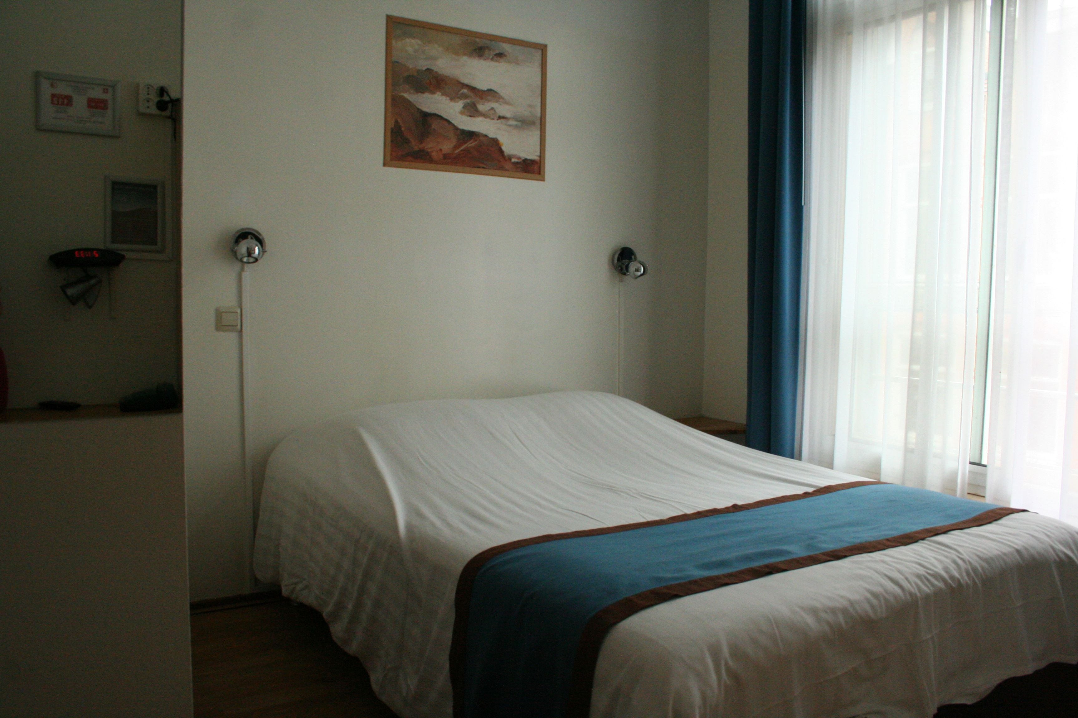 A bedroom with a bed, a painting on the wall, a window with curtains, and a wall socket.