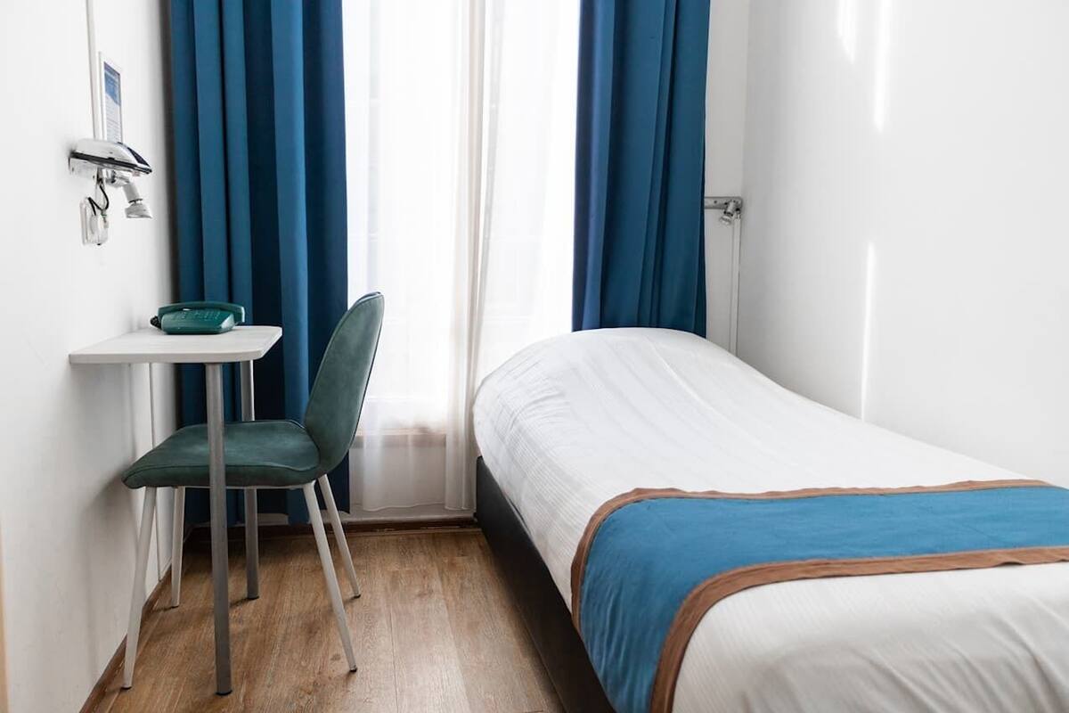 A small hotel room with a single bed, a desk with a chair, and a window with blue curtains.