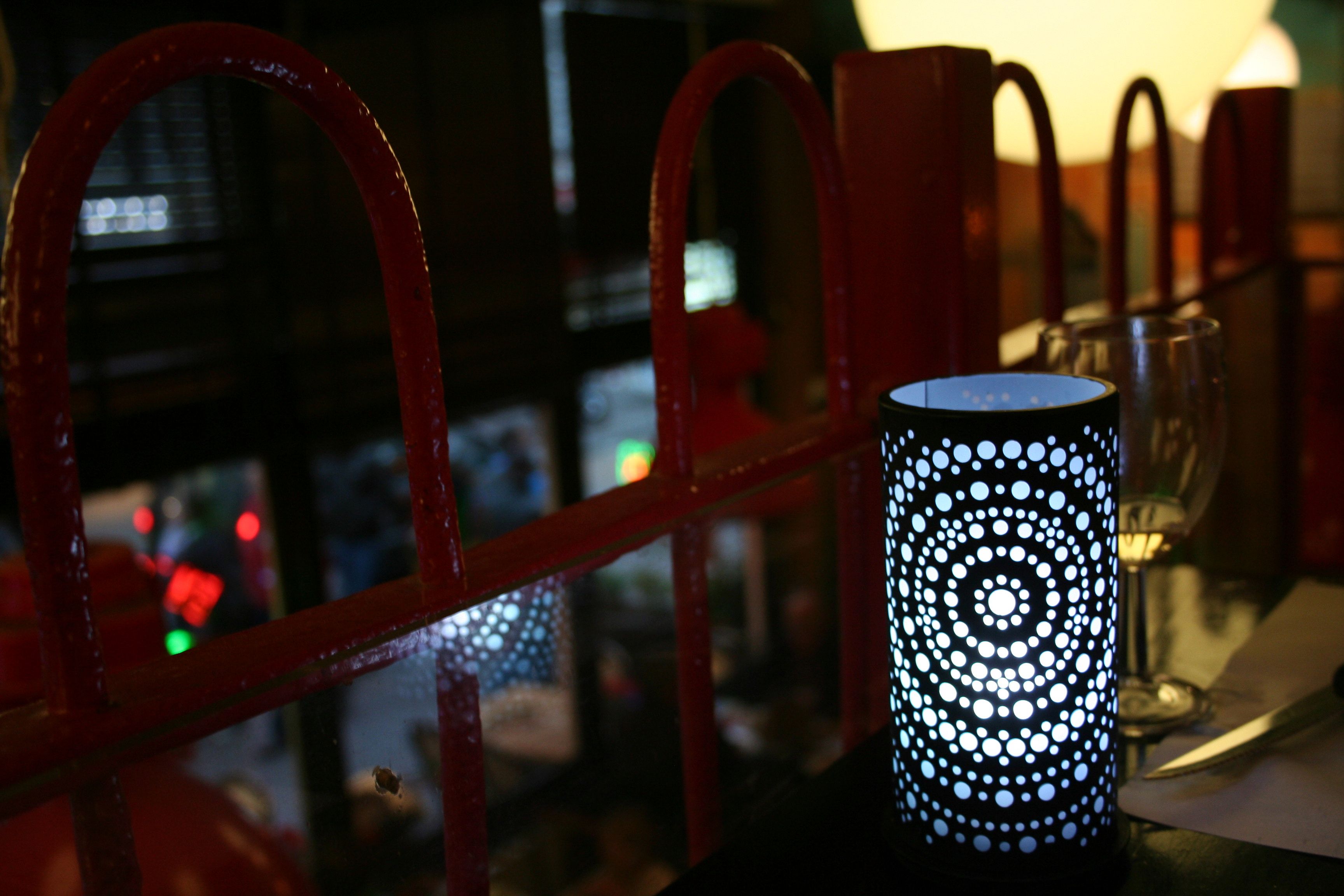 Eine zylindrische Lampe mit Muster, ein rohes Metallgeländer und ein Glastisch mit einem Weinglas.