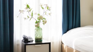 A hotel room with a bed, a nightstand with a phone, and a vase with flowers on the windowsill. - Hotel Doria (Amsterdam)