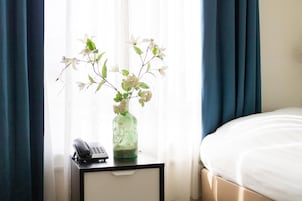 Ein Hotelzimmer mit einem Bett, einem Nachttisch mit Telefon und einer Blumenvase auf der Fensterbank.
