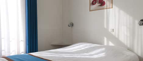 Een hotelkamer met een bed, een nachtkastje, een schilderij van appels en een raam met blauwe gordijnen.
