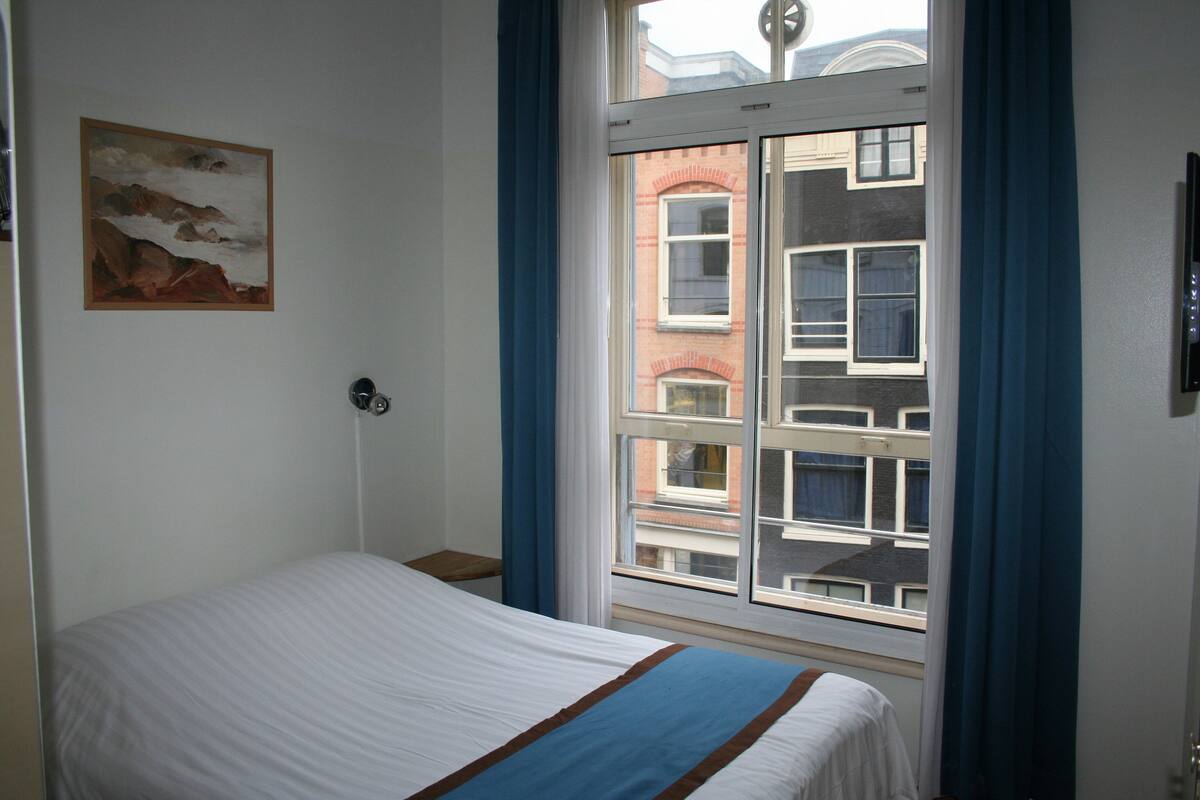 A bedroom with a bed, a window with blue curtains, and a painting on the wall.
