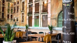 A restaurant interior with wooden tables and chairs, a menu stand labeled 'DORIA', and a view of a building with large windows and shutters. - Hotel Doria (Amsterdam)