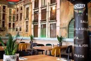 Ein Restaurantinnenraum mit Holztischen und -stühlen, einem Menüständer mit der Aufschrift 'DORIA' und Blick auf ein Gebäude mit großen Fenstern und Fensterläden.