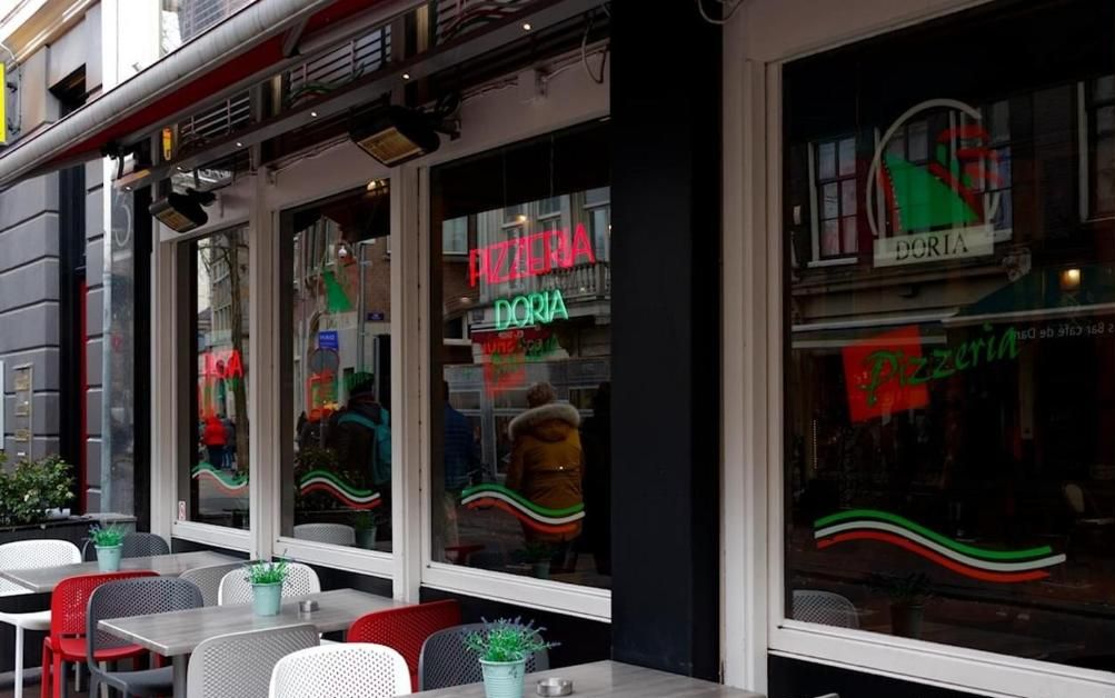 An outdoor seating area with tables and chairs, a neon sign for a pizzeria, and a window with a reflection of a person.