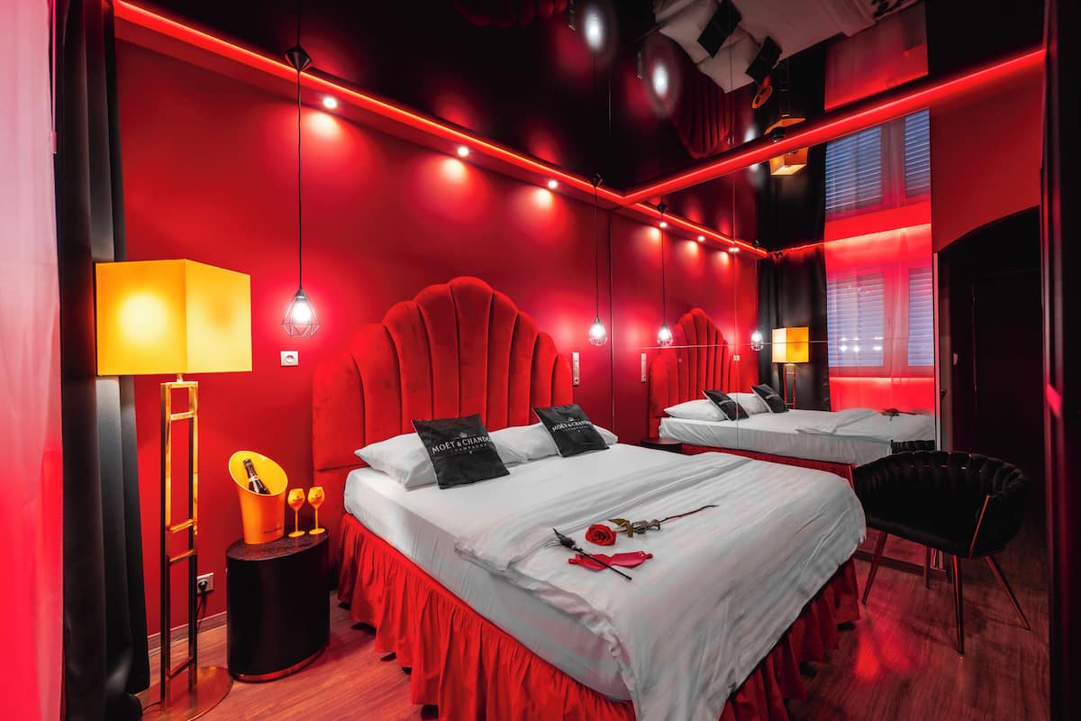 A red-themed hotel room with a large bed, a side table with a lamp, and a mirror reflecting the room.