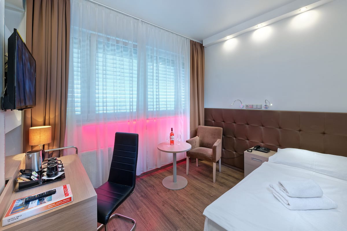 A hotel room with a bed, a desk with a lamp, a chair, a small table with a bottle and glasses, and a TV on the wall.