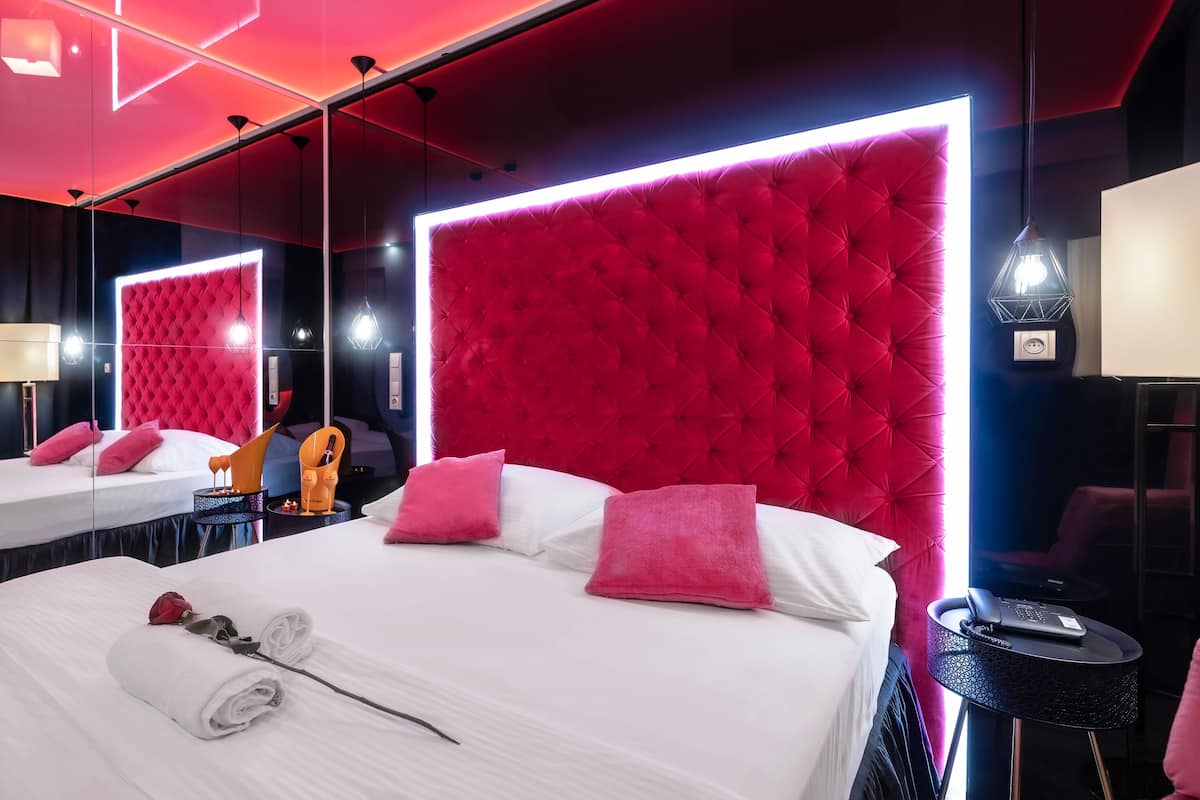 A hotel room with two beds, each with white linens and pink pillows, a red tufted headboard, and a small table with a lamp.