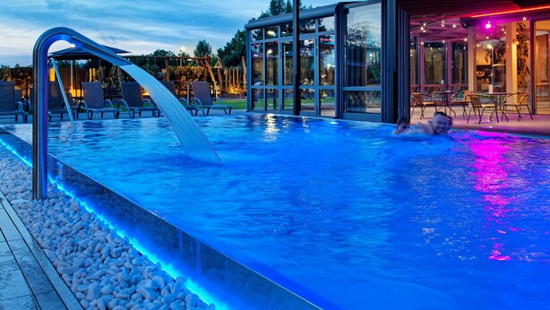 En swimmingpool med et glasmur, et vandfald og en terrasse med stole og borde.