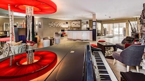Un salon moderne avec un piano, une structure circulaire rouge et un coin cuisine à l’arrière-plan.