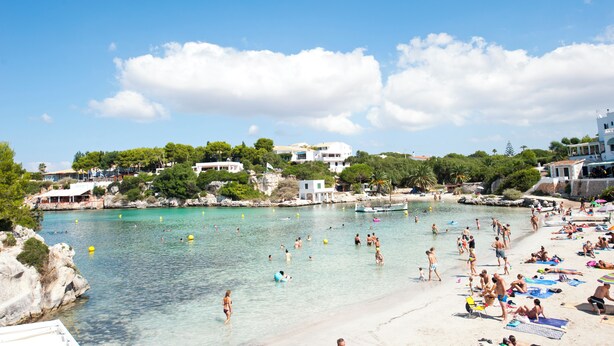 Una playa con aguas turquesas cristalinas, gente nadando y tomando el sol, y edificios al fondo.