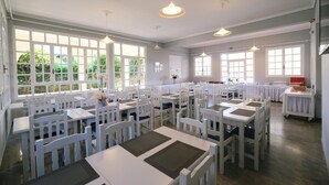 Un comedor amplio con mesas y sillas blancas, ventanas grandes e iluminación colgante.