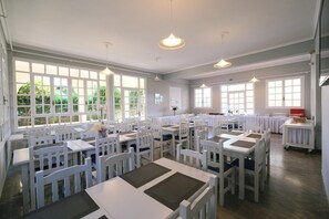 A spacious dining area with white tables and chairs, large windows, and pendant lighting. - Ifestos Hotel (MIRINA)