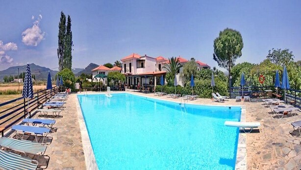A swimming pool with lounge chairs and umbrellas, a building with red roofs, and a mountainous backdrop. - Pela Hotel (Lesvos)