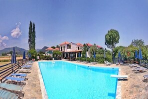 A swimming pool with lounge chairs and umbrellas, a building with red roofs, and a mountainous backdrop. - Pela Hotel (Lesvos)