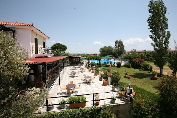 A patio area with outdoor seating and a pool in the background. - Pela Hotel (Lesvos)