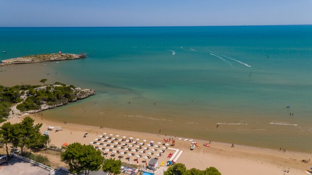 A beach with clear turquoise water, a sandy shore, and several umbrellas and lounge chairs. - Villaggio Capo Vieste Camping (Vieste)