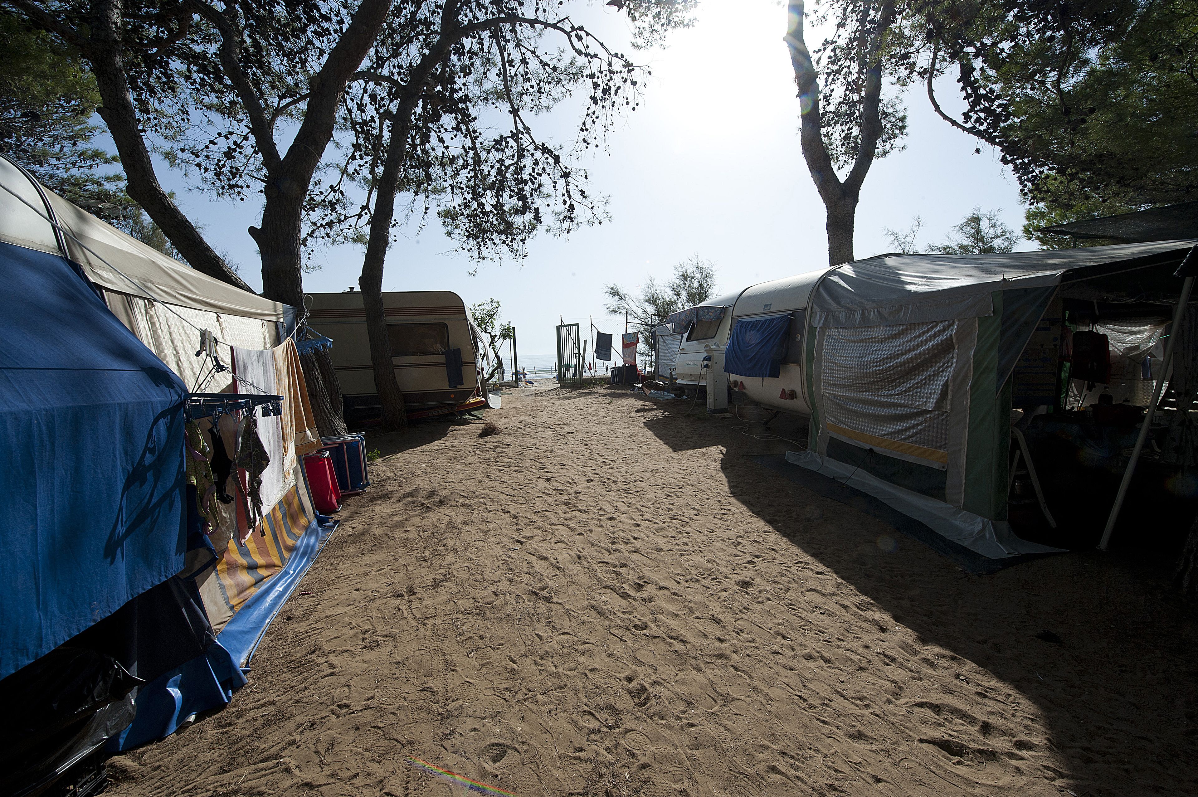 Un camping con varias caravanas y tiendas de campaña, suelo arenoso y árboles que dan sombra.