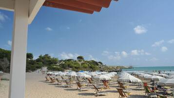 Une plage bordée de rangées de chaises de plage et de parasols, un ciel dégagé et, au loin, une colline rocheuse.