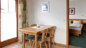 A room with a wooden dining table and chairs, a bed with a wooden headboard, and a window with curtains. - Alpenland (FLACHAU)