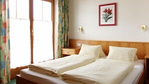 A double bed with white linens, a wooden headboard, a nightstand, a wall-mounted lamp, and a framed floral artwork. - Alpenland (FLACHAU)