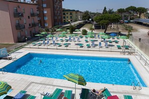 A swimming pool with lounge chairs and umbrellas, surrounded by buildings and trees. - Hotel Vianello (Jesolo)