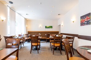 Un restaurant avec des tables et des chaises en bois, un plafond haut doté d’un éclairage encastré et un tableau accroché au mur.