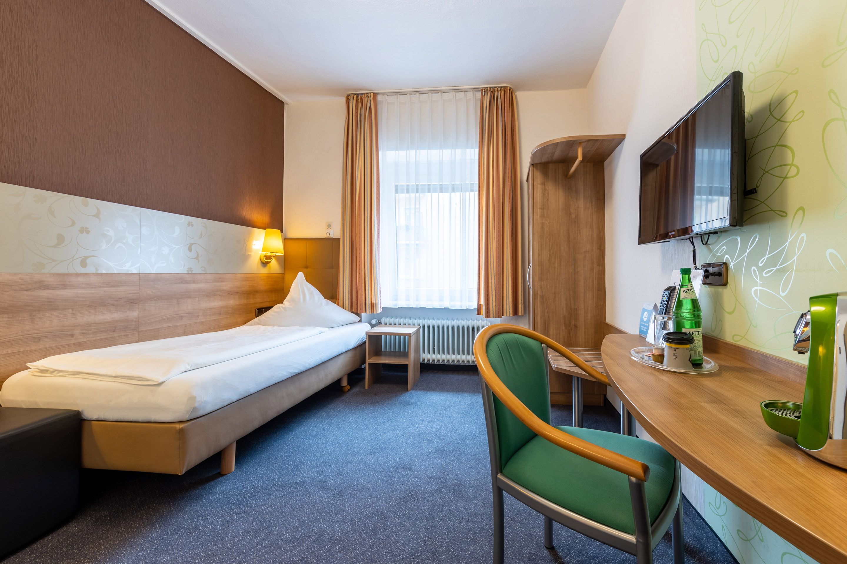 A hotel room with a bed, a desk with a chair, a television, and a window with curtains.
