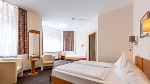 Ein Hotelzimmer mit einem großen Bett, zwei Stühlen, einem kleinen Tisch, einem Spiegel und einem Fenster mit Vorhängen.