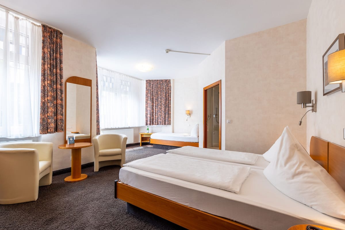 Habitación de hotel con una cama grande, dos sillas, una mesita, un espejo y una ventana con cortinas.