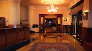 A lobby with a reception desk, a chandelier, a patterned carpet, and a red leather sofa. - Principe Palace (Jesolo)