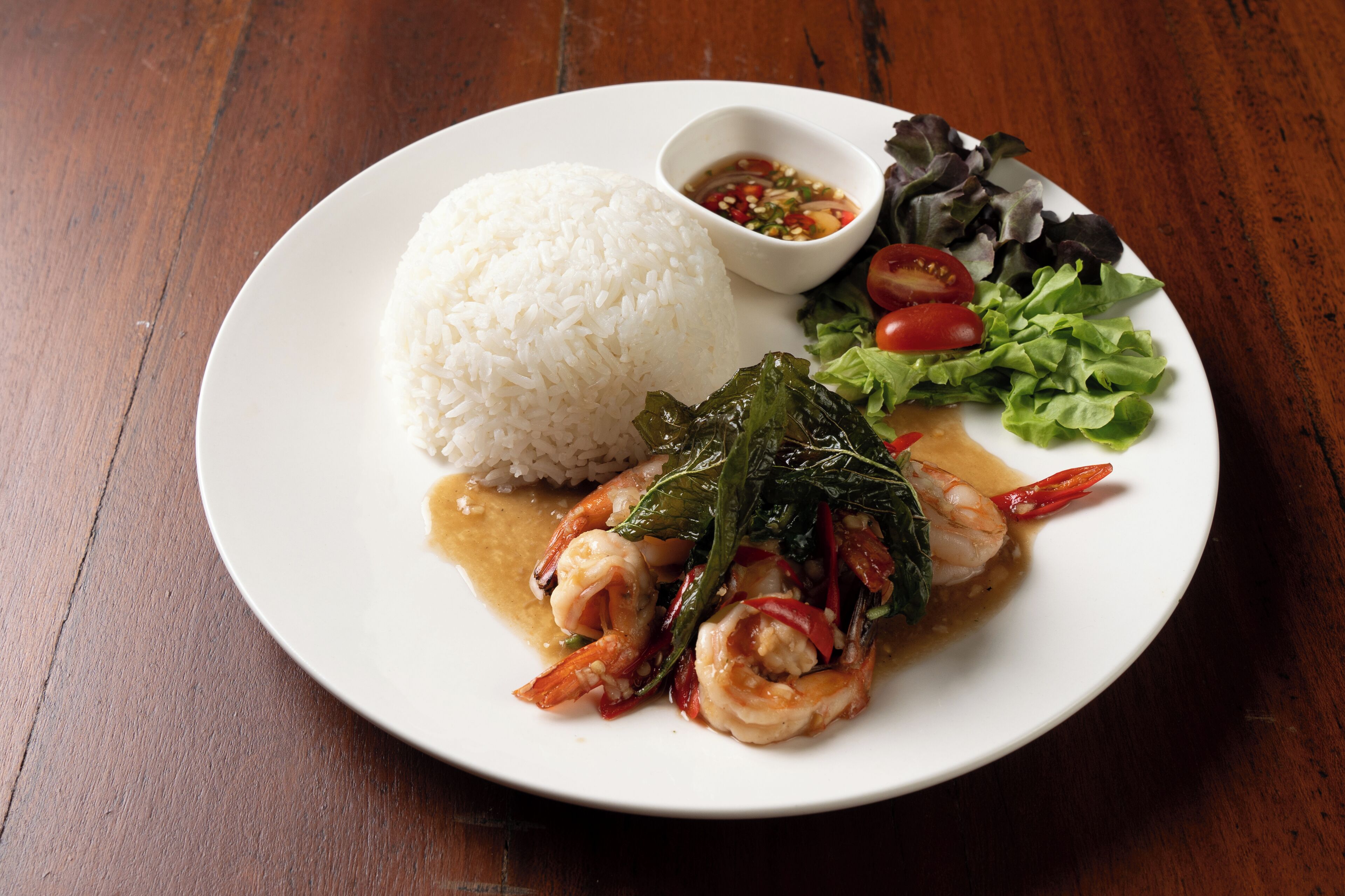 A plate of white rice, shrimp, vegetables, and a small bowl of sauce on a wooden table.
