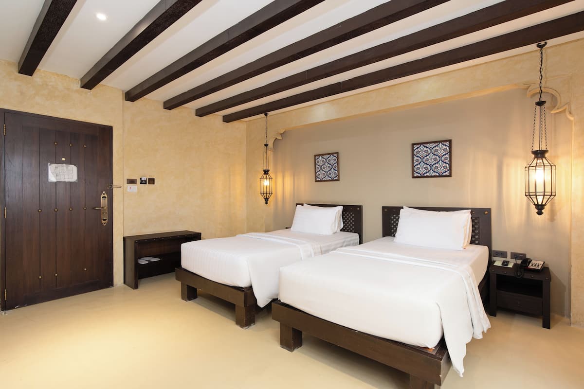 A hotel room with two beds, wooden headboards, a dark wooden door, a small bedside table with a phone and a clock, and a wall-mounted light fixture.