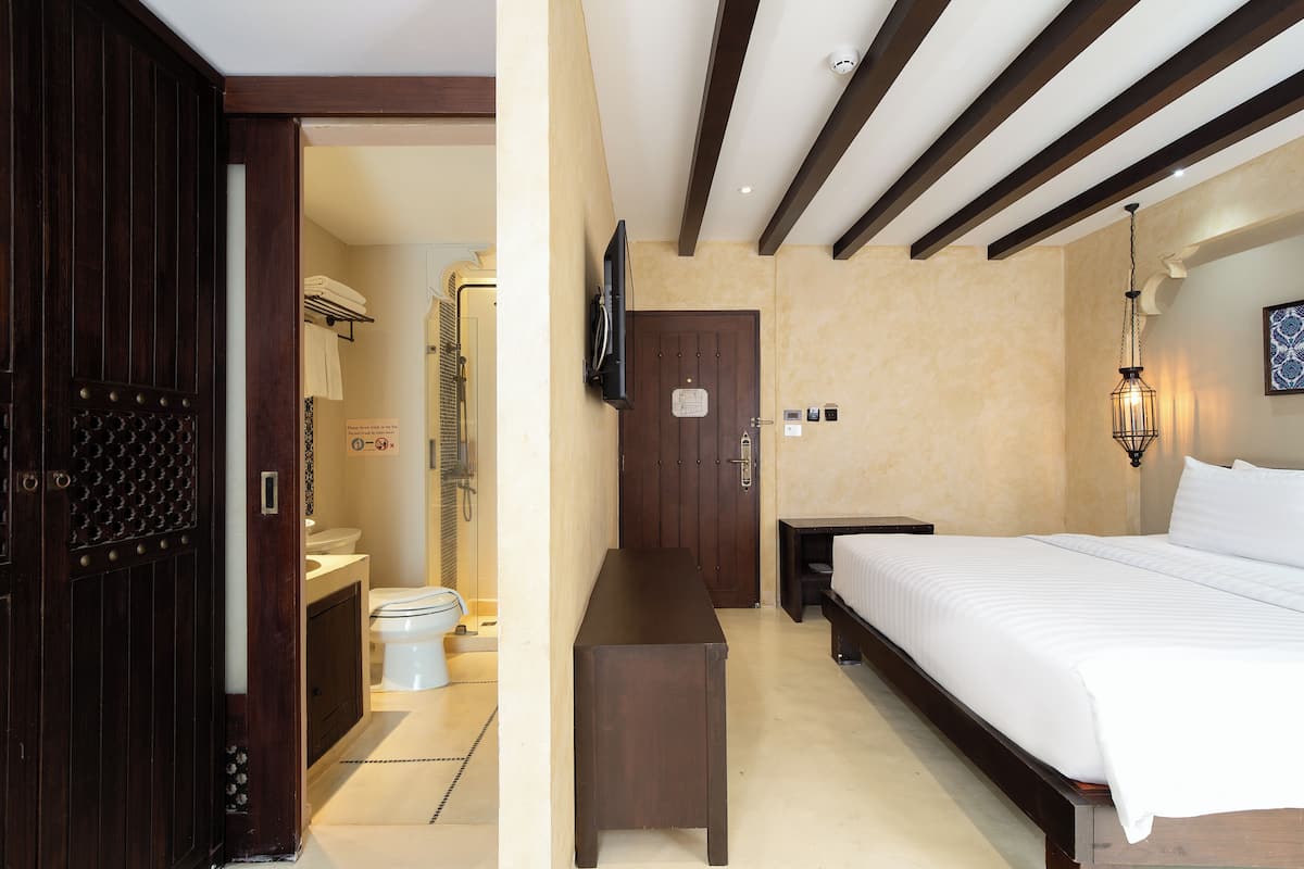A hotel room with a wooden door, a bed with white bedding, a small table, a wall-mounted TV, and a bathroom visible through an open door.